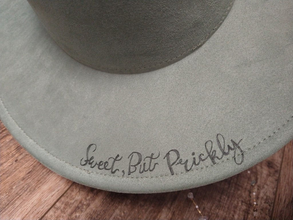Gray hat with 'Sweet, But Prickly' embroidery on a wooden surface