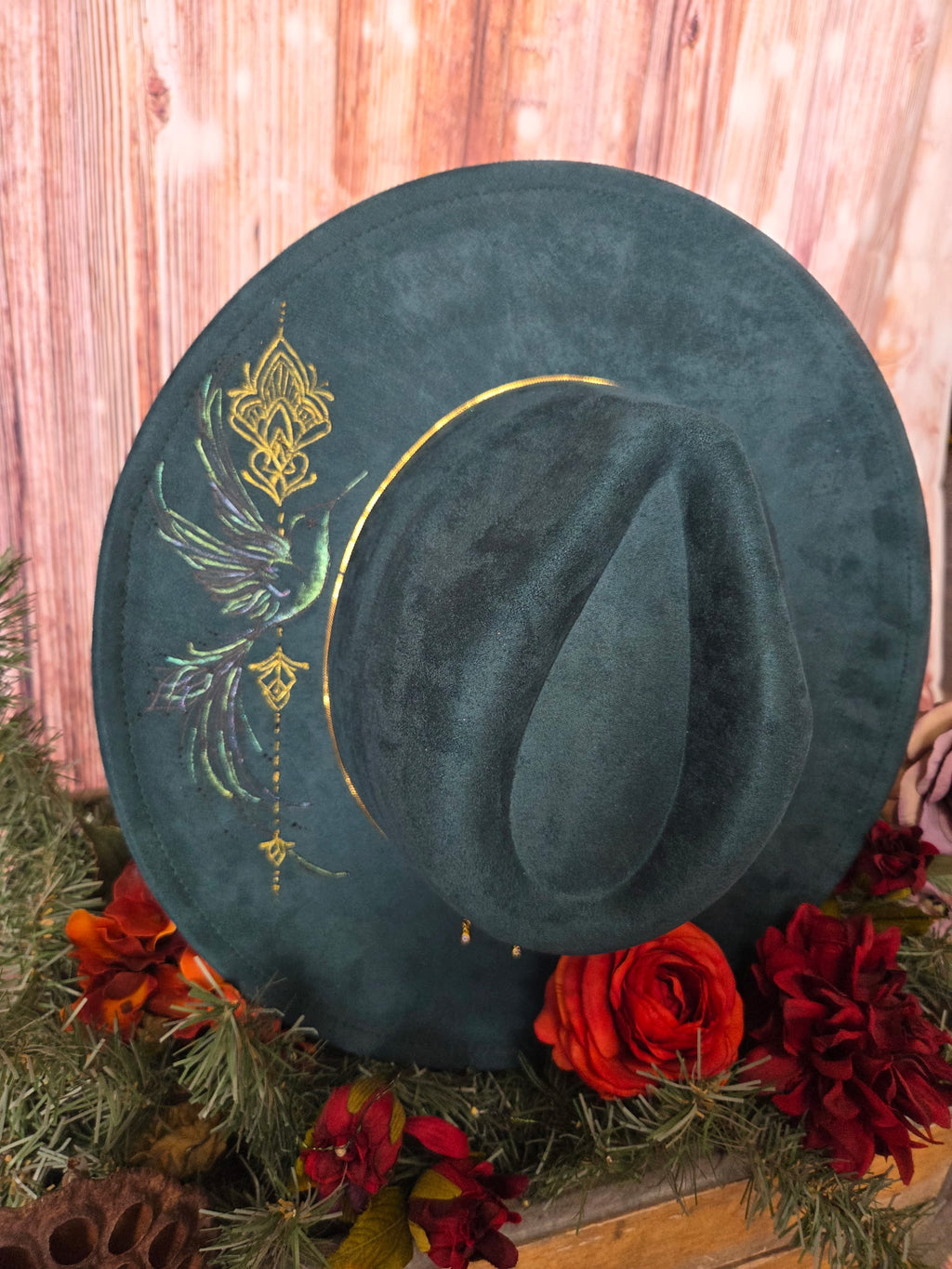Green velvet hat with gold paint on a decorative background with red flowers and greenery.