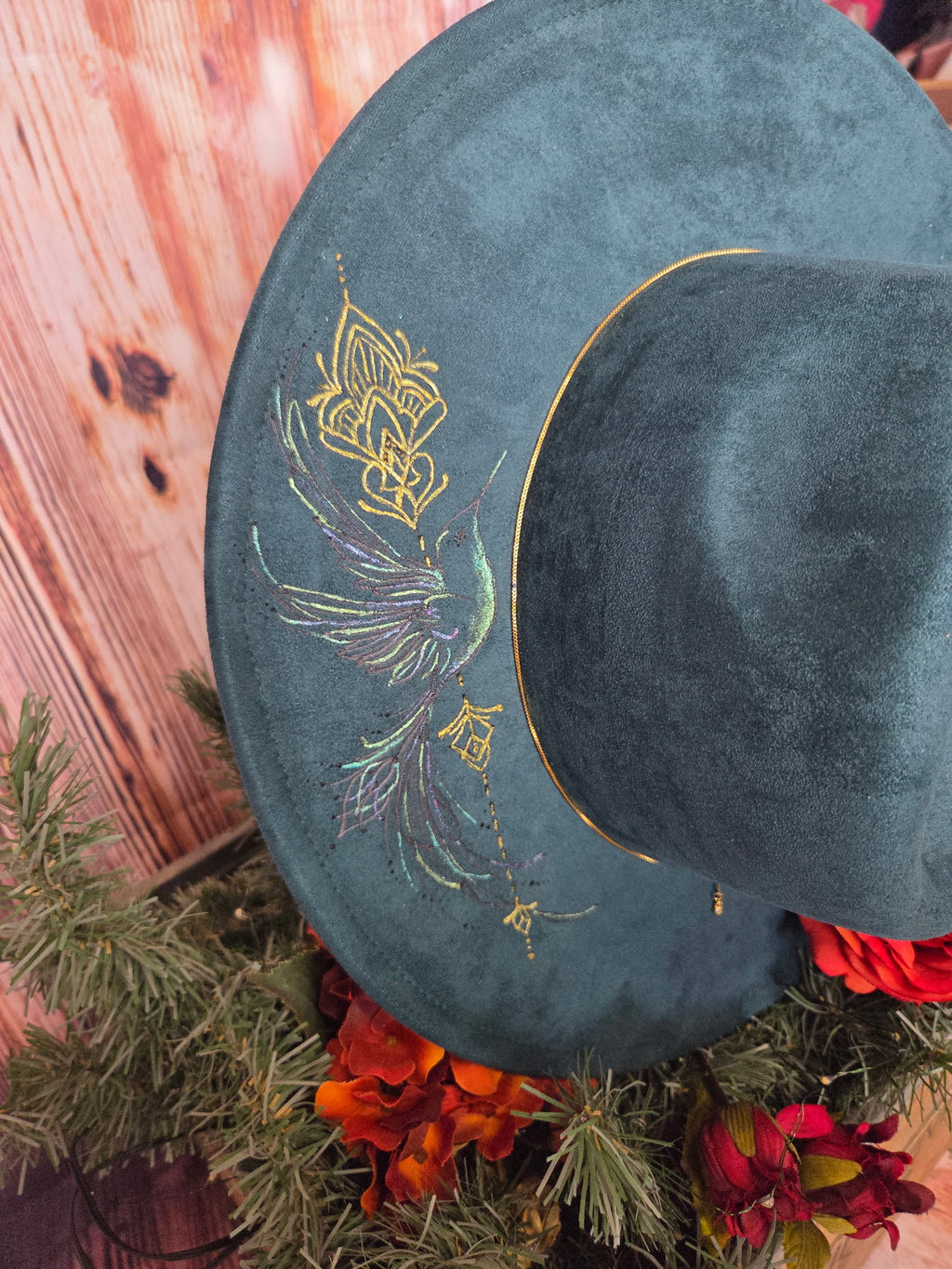 Dark green hat with gold on a wooden surface with greenery and red flowers.
