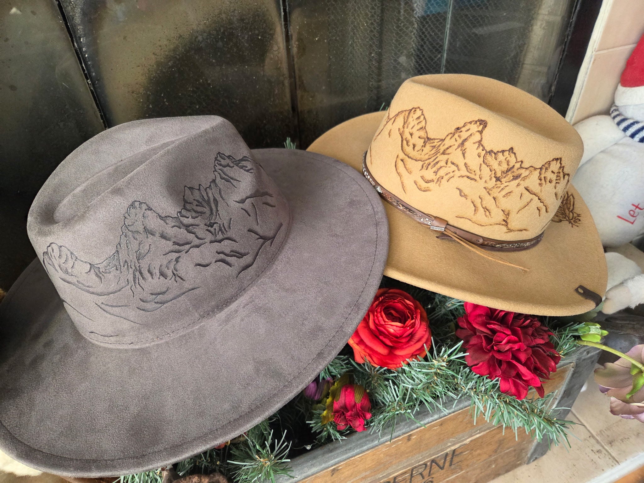 Two cowboy hats, one gray and one brown, with floral decorations on a metal surface.